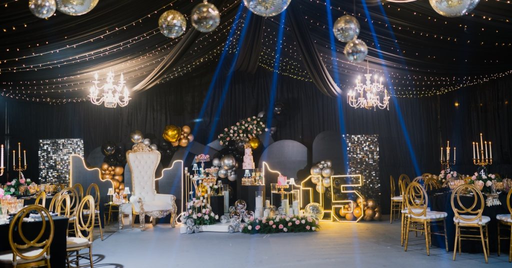 Black-draped party venue features mirrored disco balls, blue spotlights, gold seating, flowers, and a glowing 15.